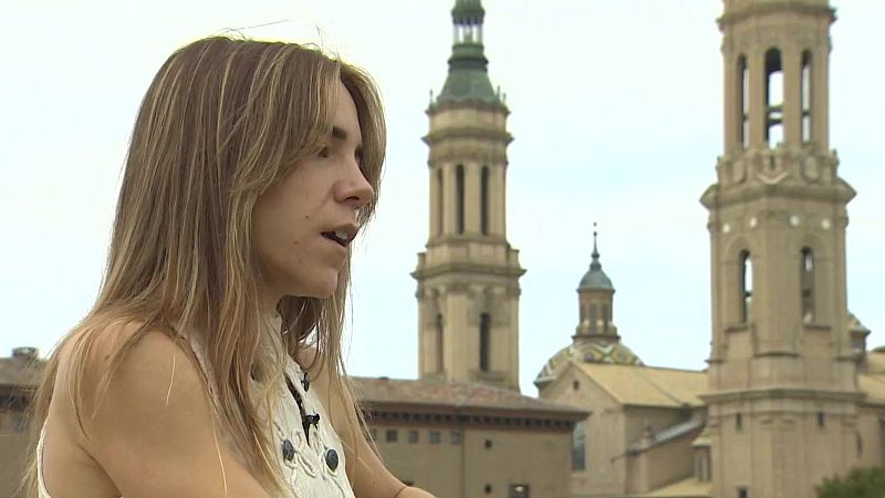 Mujer de perfil hablando en exteriores con la Basílica del Pilar de Zaragoza al fondo bajo un cielo nublado.