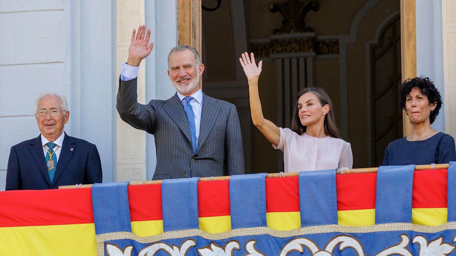 Los Reyes de España saludan desde el balcón de un edificio decorado en un acto oficial.