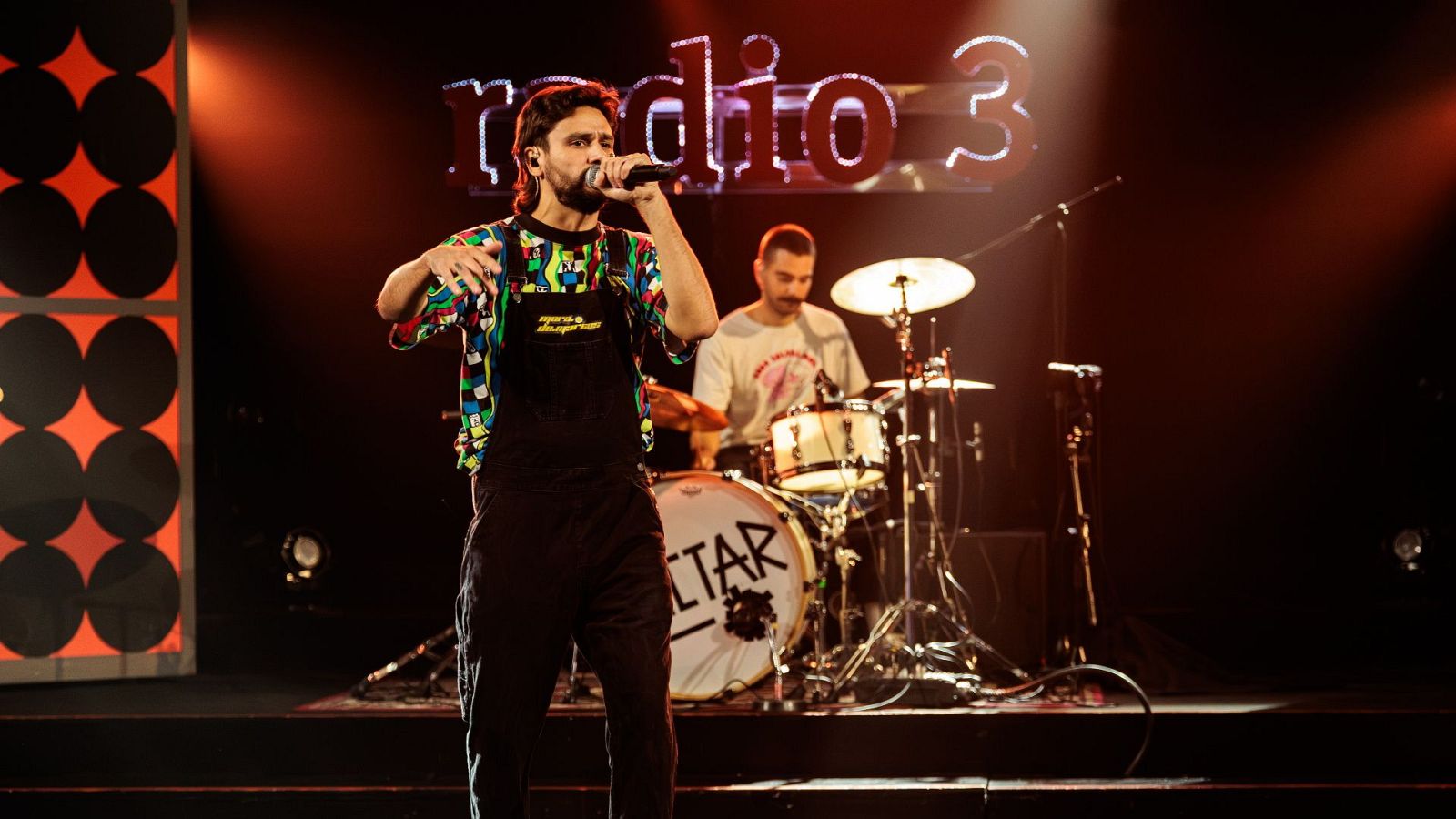 Un cantante con peto negro y camiseta estampada actúa en directo junto a su banda bajo una iluminación rojiza y el rótulo de Radio 3.