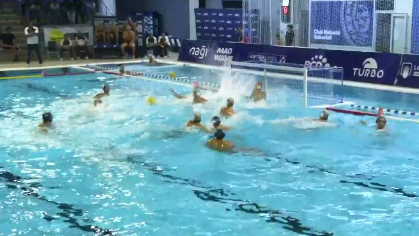 Jugadores de waterpolo compiten en una piscina cubierta rodeados de salpicaduras y con una portería flotante y un marcador electrónico de fondo.