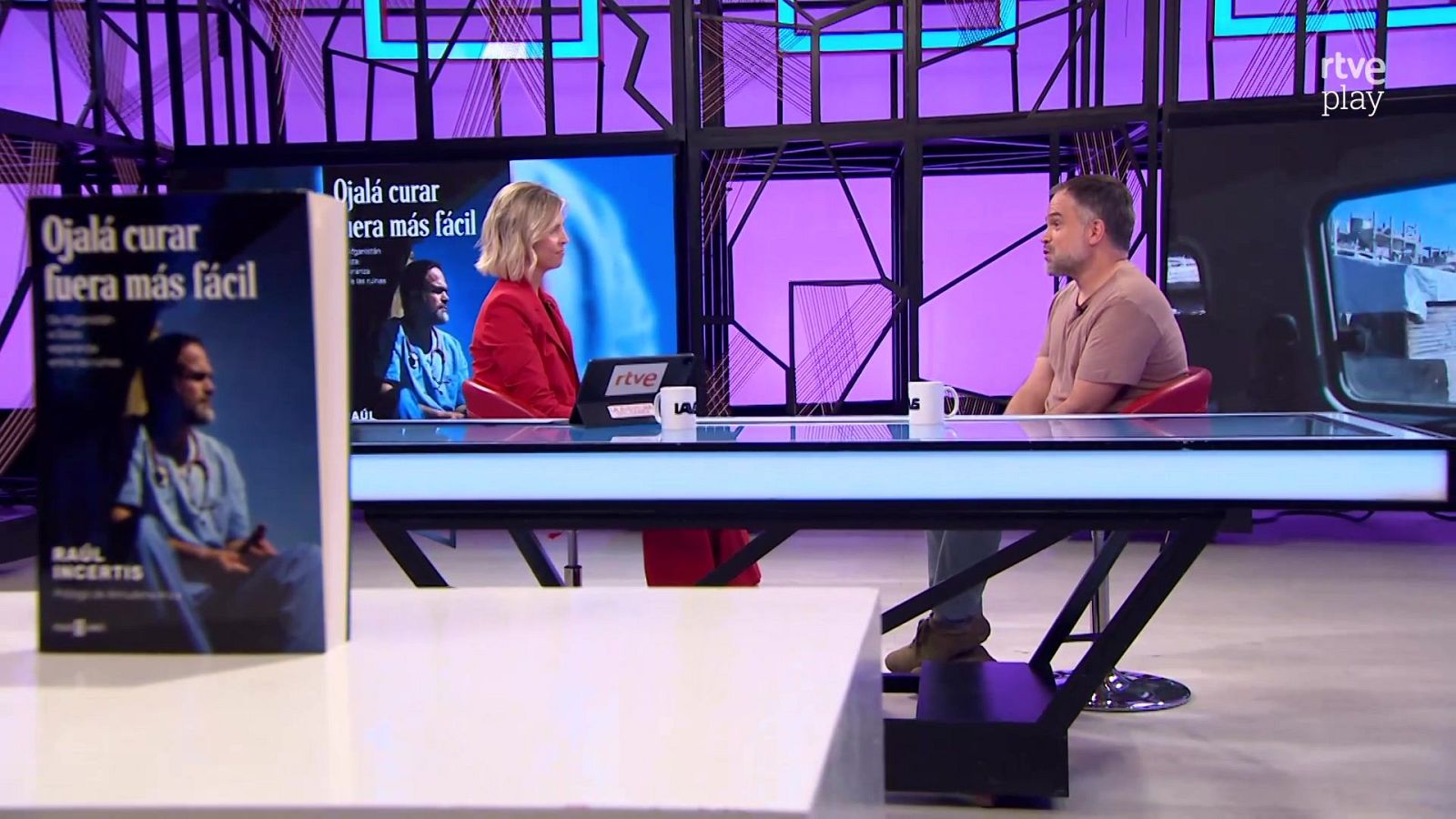 Mara Peterssen entrevista al médico Raúl Incertis en un plató moderno iluminado en tonos azules y fucsias, con un ejemplar de su libro sobre la mesa.