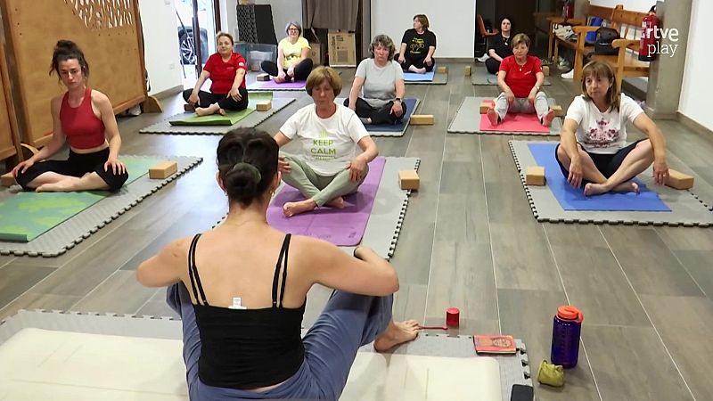 Clase de yoga en sala interior con instructora de espaldas y varias alumnas sentadas en esterillas de colores sobre suelo de gomaespuma.