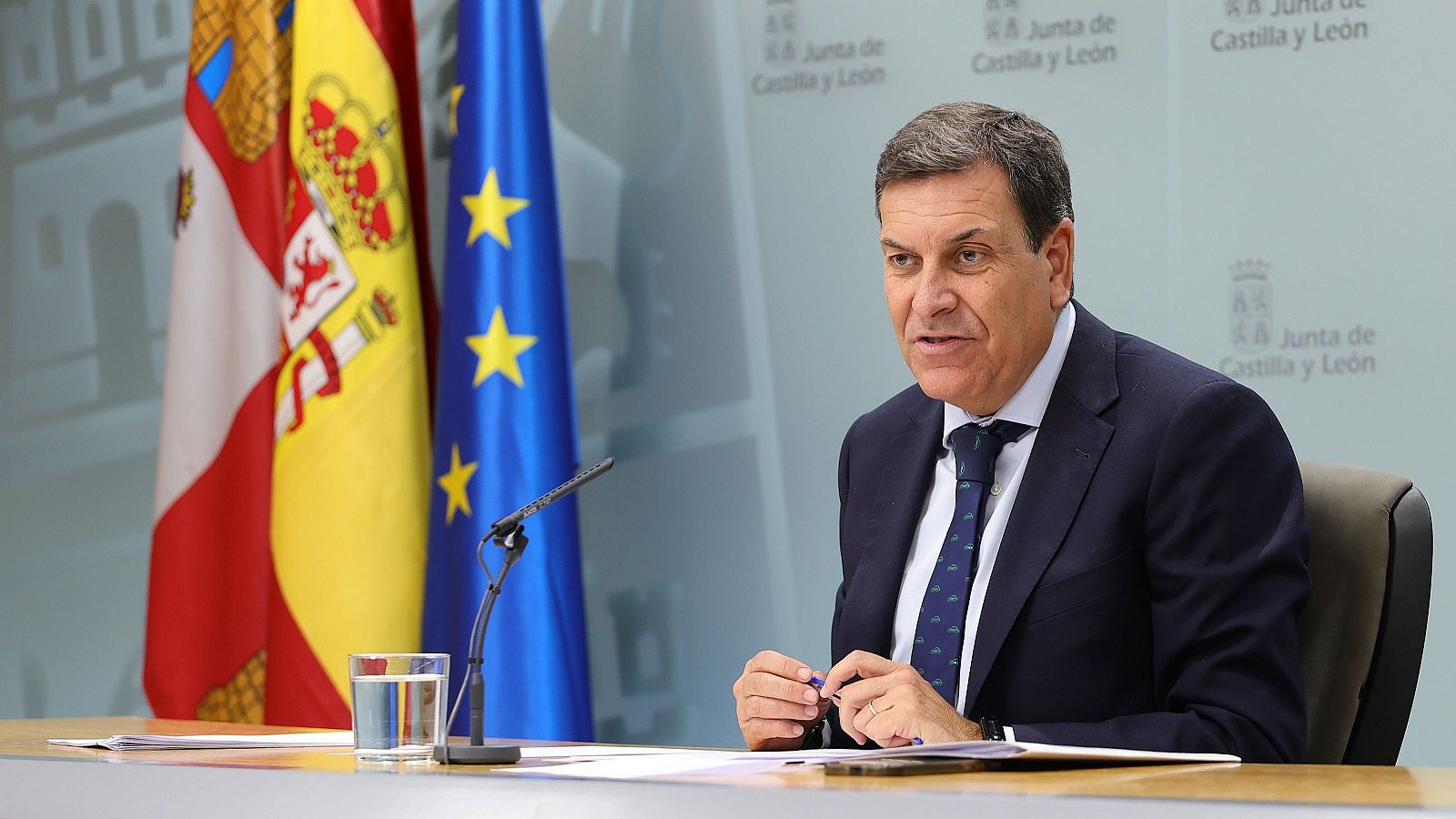 Carlos Fernández Carriedo ofrece una rueda de prensa sentado ante una mesa con documentos y un micrófono, con traje oscuro, corbata y las banderas oficiales al fondo.