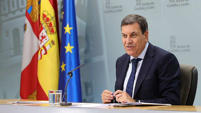 Carlos Fernández Carriedo ofrece una rueda de prensa sentado ante una mesa con documentos y un micrófono, con traje oscuro, corbata y las banderas oficiales al fondo.