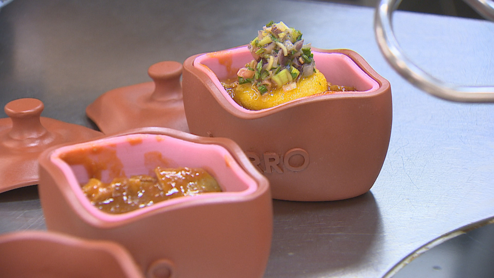 Pequeñas cazuelas cuadradas de barro sobre encimera de acero inoxidable con un guiso tradicional y verduras.