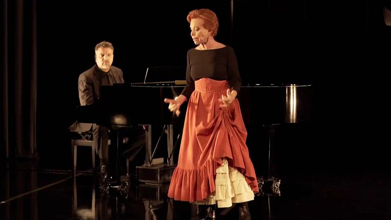 Intérprete con cabello rojizo y falda larga terracota cantando en un escenario junto a un pianista con piano de cola.
