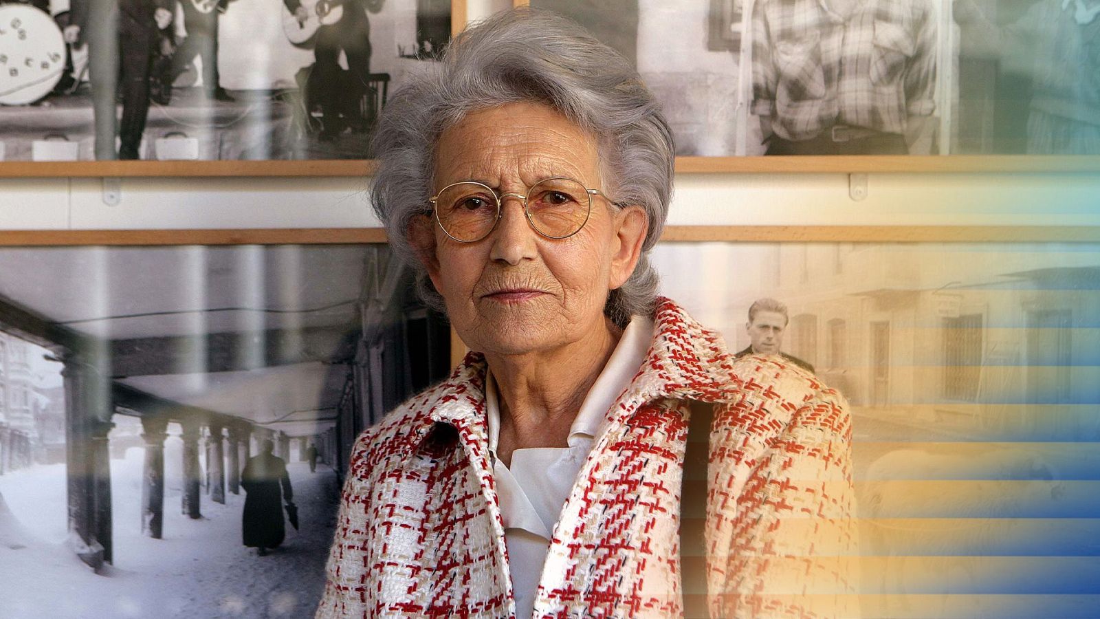 La fotógrafa Piedad Isla, con cabello canoso y gafas de montura fina, posa seria frente a una exposición de sus fotografías en blanco y negro sobre la vida rural.