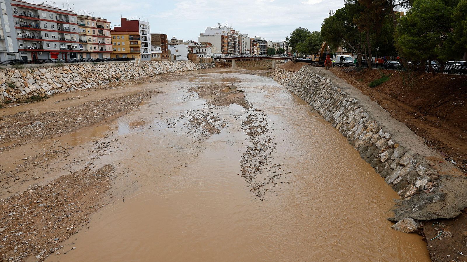 Cauce fluvial canalizado con agua fangosa, flanqueado por edificios, un puente al fondo y una excavadora trabajando en la orilla bajo un cielo nublado.