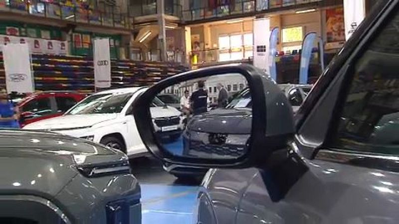 Interior de un salón del automóvil con varios vehículos en exposición, visitantes y carteles publicitarios de marcas en un pabellón con gradas.