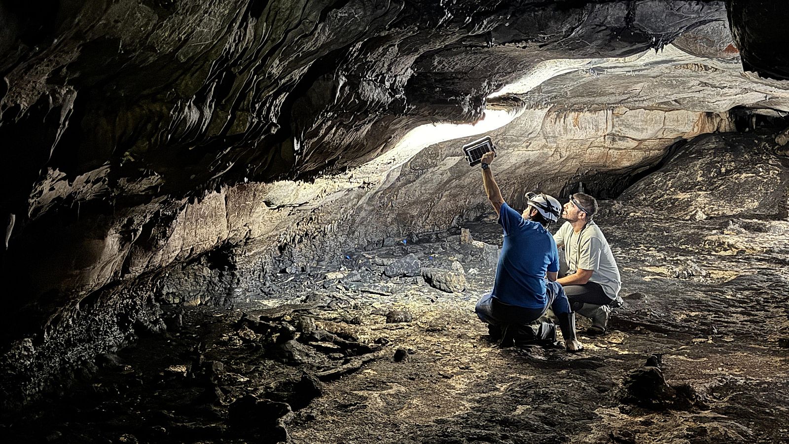 Dos investigadores con equipamiento de montaña examinan el interior de una cueva subterránea, utilizando una tableta digital para escanear la bóveda rocosa iluminada.