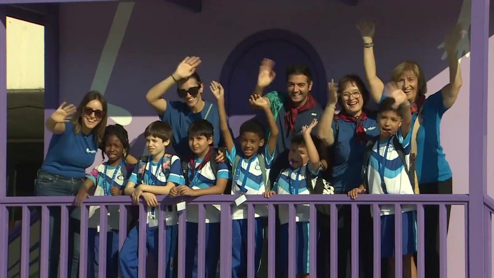 Grupo de once personas, entre niños y adultos sonrientes, saludan desde el porche de una estructura de madera morada con camisetas deportivas y acreditaciones.
