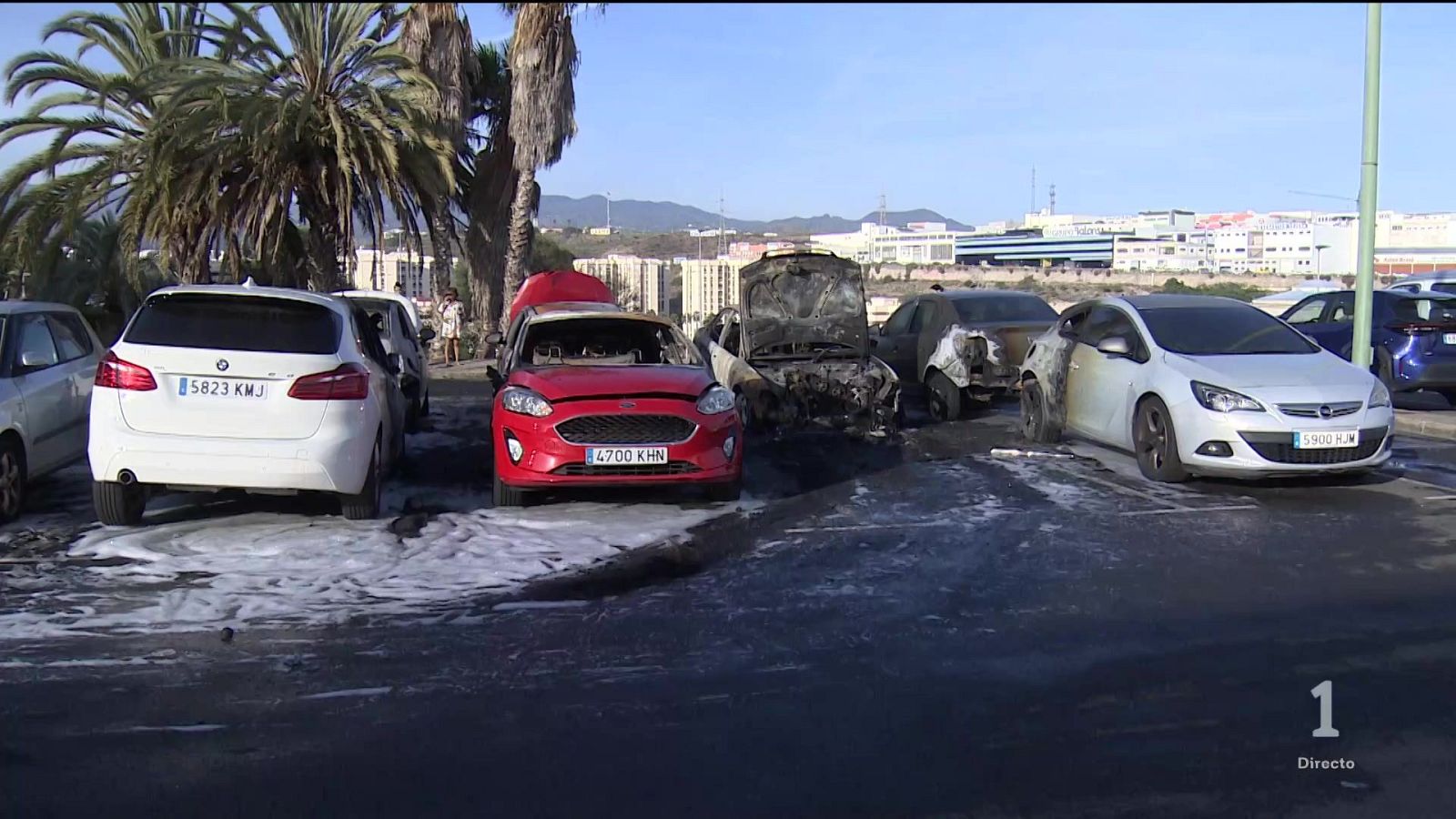 Vehículos calcinados y dañados por un incendio en un aparcamiento con espuma extintora en el asfalto.
