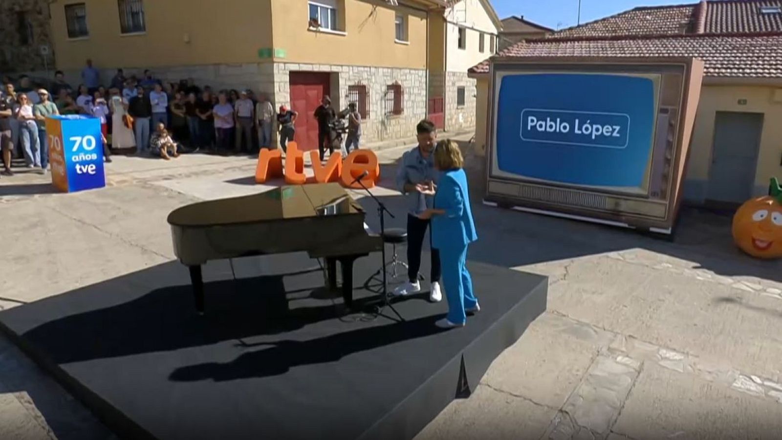 Pablo López conversa junto a un piano en una plaza decorada con elementos de RTVE ante la mirada de varios vecinos.