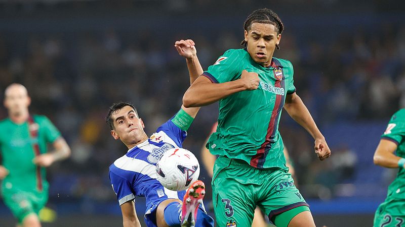 Intensa disputa por el bal�n entre un futbolista con equipaci�n verde azulada y otro con camiseta blanquiazul que se desliza para despejar el esf�rico.