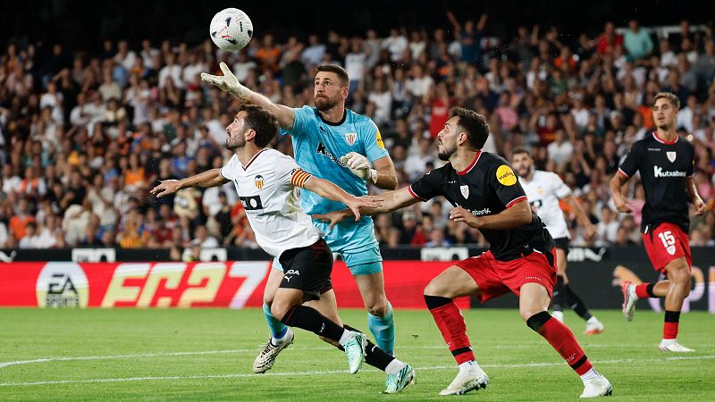 Partido de f�tbol nocturno con varios jugadores disputando un bal�n a�reo y el guardameta despejando con el pu�o en un estadio lleno.