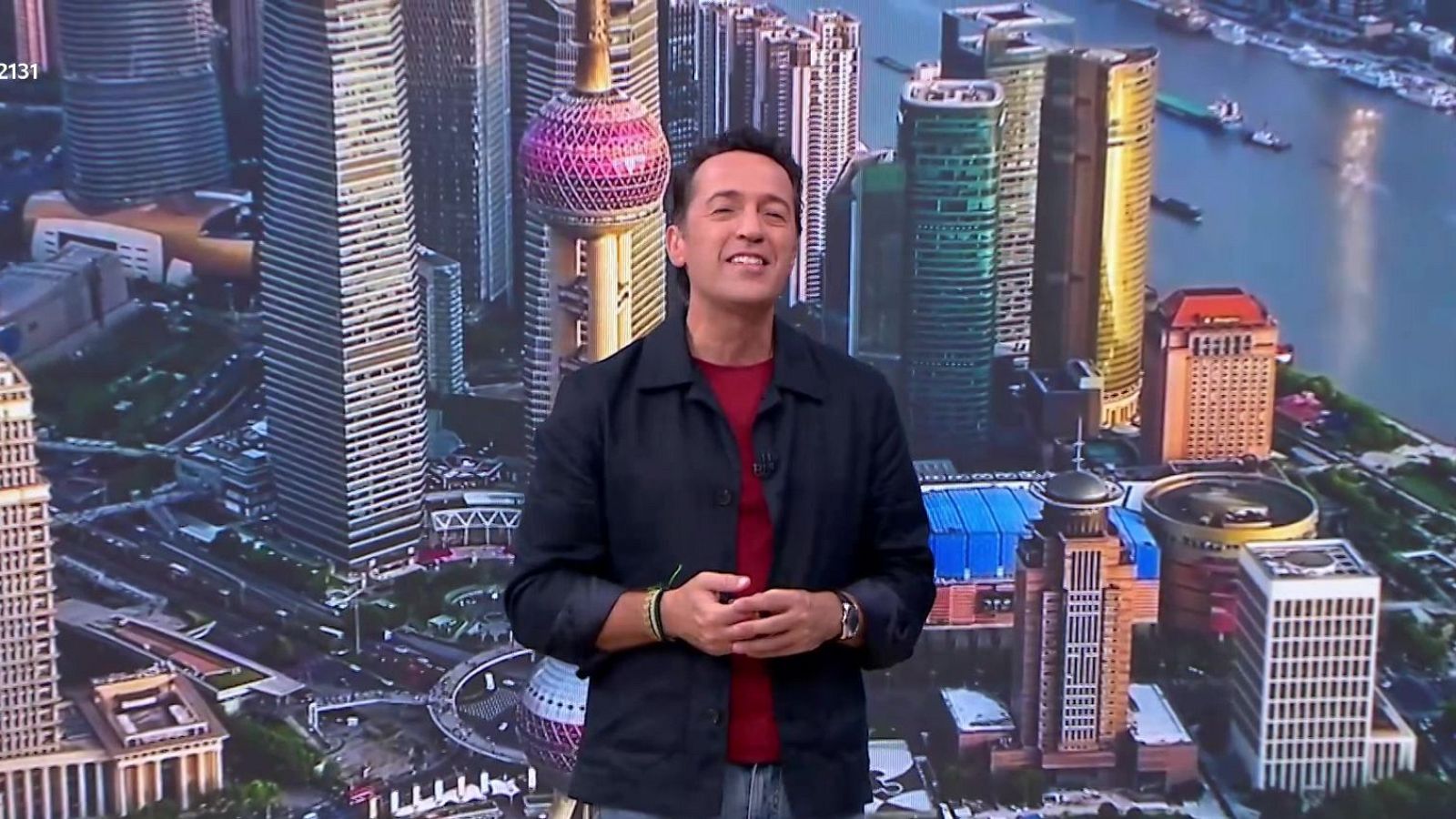 El geógrafo y presentador Jacob Petrus sonríe a cámara en el plató frente a una pantalla con una vista aérea de Shanghái y la Torre de la Perla Oriental.