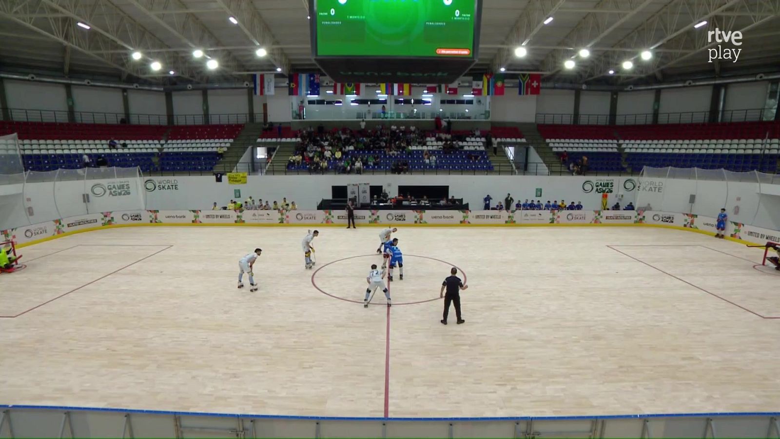 Partido de hockey sobre patines en un pabellón cubierto con pista de madera, jugadores en el centro y marcador electrónico al fondo.
