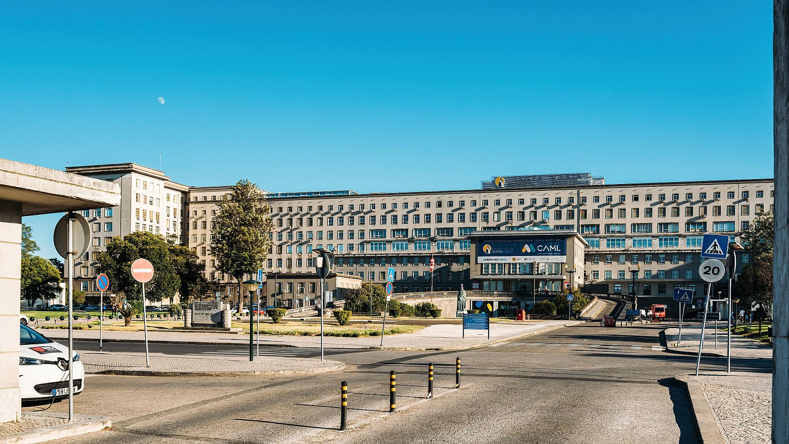 Vista exterior del Hospital de Santa María en Lisboa con su fachada racionalista, rodeado de zonas verdes, calzadas y señales de tráfico bajo un cielo azul.