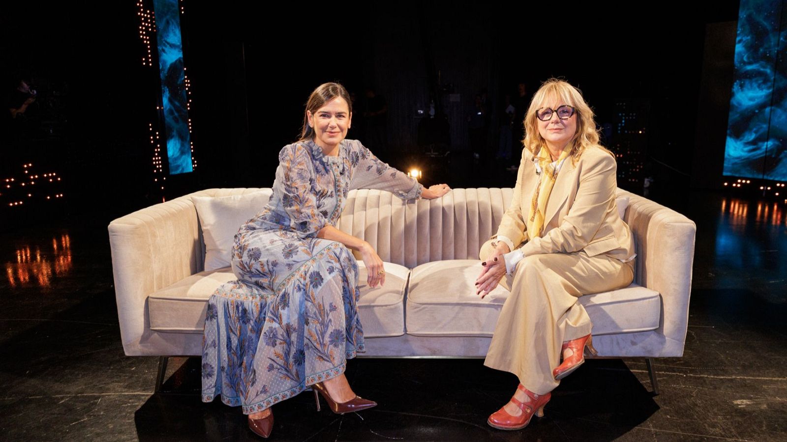 La presentadora Elena Sánchez y la actriz Emma Suárez posan sonrientes en un sofá de terciopelo beige en un plató de televisión con paneles luminosos de fondo.