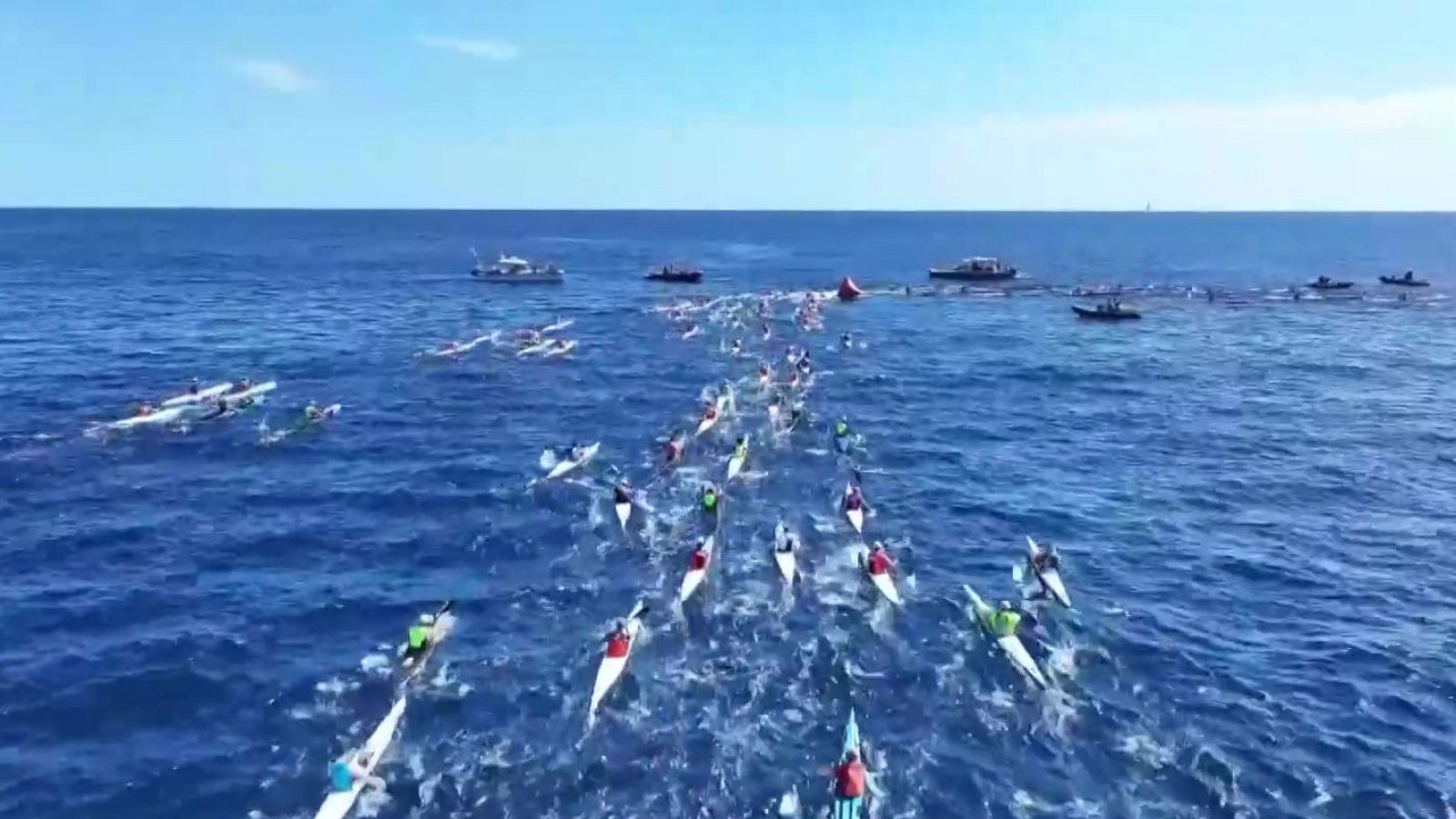 Vista aérea de una regata de piragüismo en mar abierto con numerosos kayaks avanzando hacia una boya roja bajo un cielo despejado.
