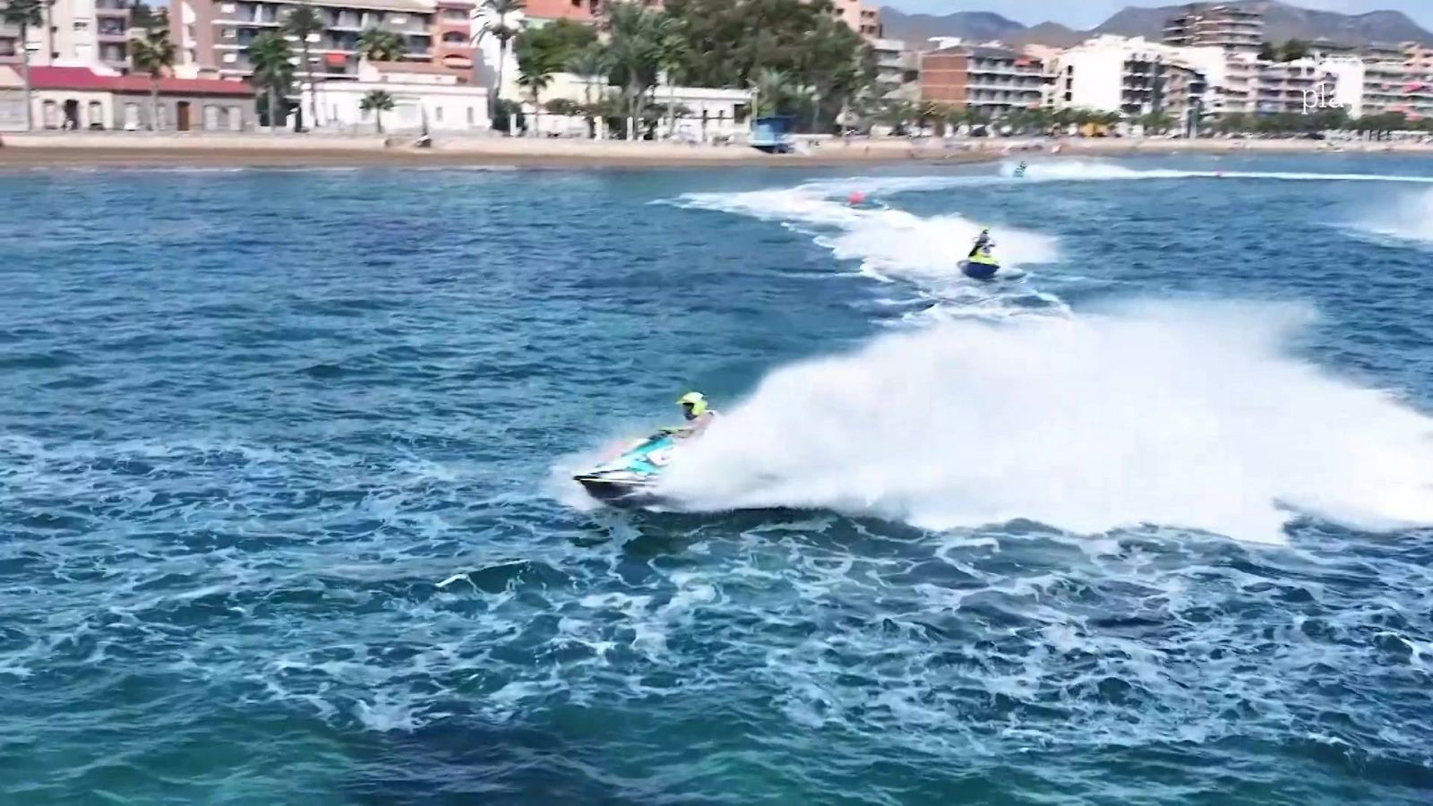 Piloto de moto acuática con casco amarillo realiza un giro pronunciado en el mar, levantando una gran cortina de espuma cerca de la costa con edificios y palmeras.
