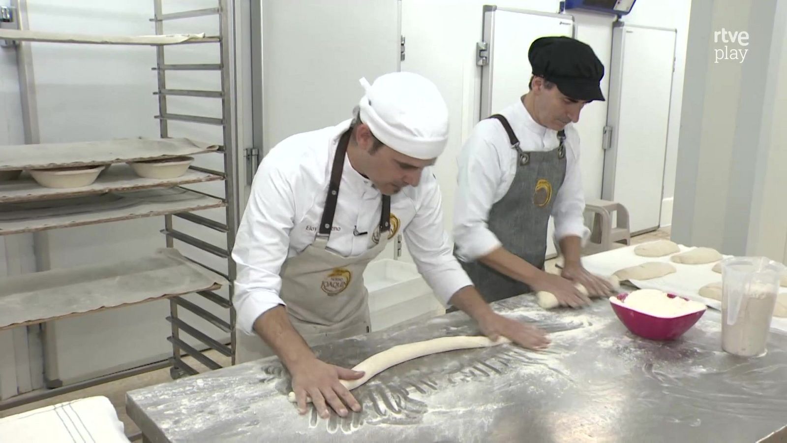 Dos panaderos con chaquetillas y delantales trabajan en una mesa de acero inoxidable moldeando masa en un obrador profesional con un logotipo televisivo.