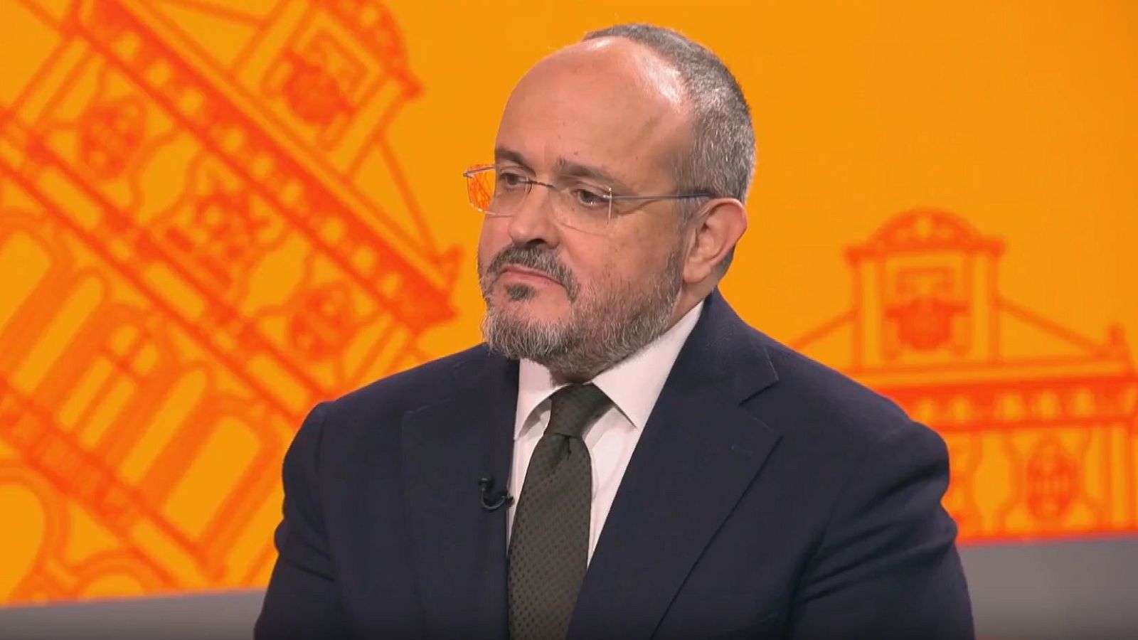 Alejandro Fernández, político con gafas y barba, viste traje y corbata en un plató de televisión con fondo naranja.