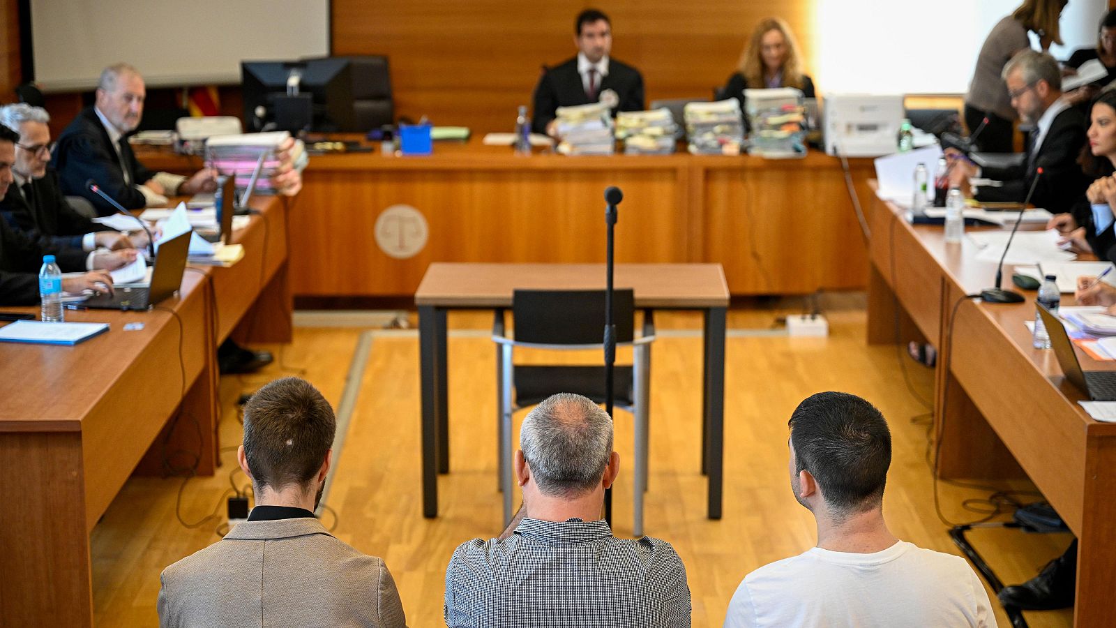 Sala judicial durante una vista oral con tres hombres en el banquillo en primer plano, letrados con toga en los laterales y el juez al fondo sobre tarima elevada.