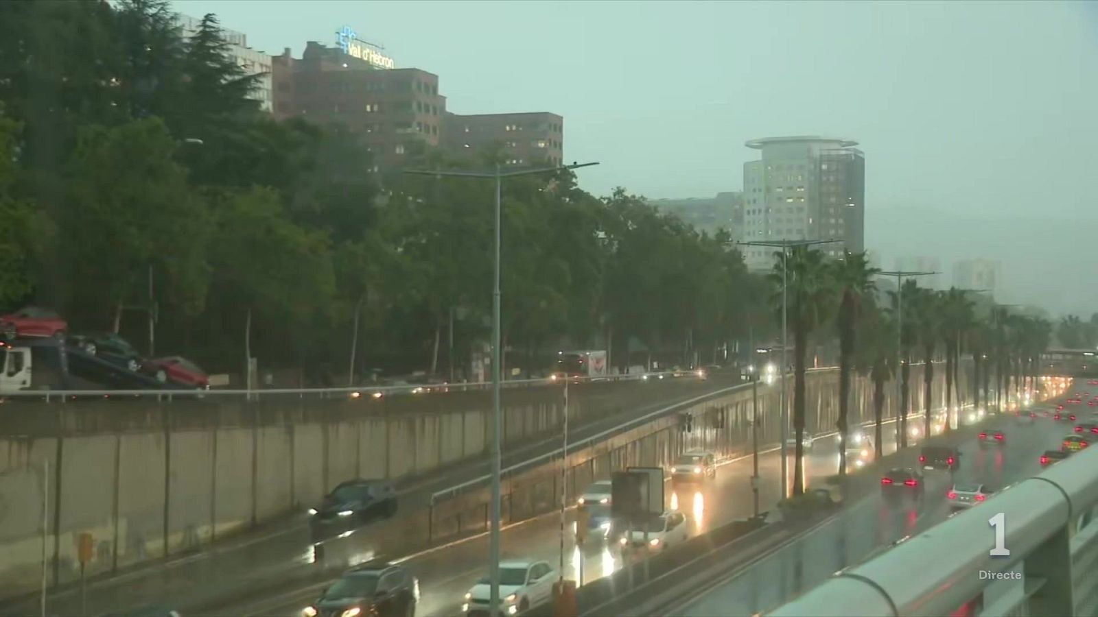 Plano picado de tráfico en autovía urbana bajo lluvia intensa con asfalto mojado y luminoso reflejo de faros, con edificios al fondo y logotipo de 'La 1' en pantalla.