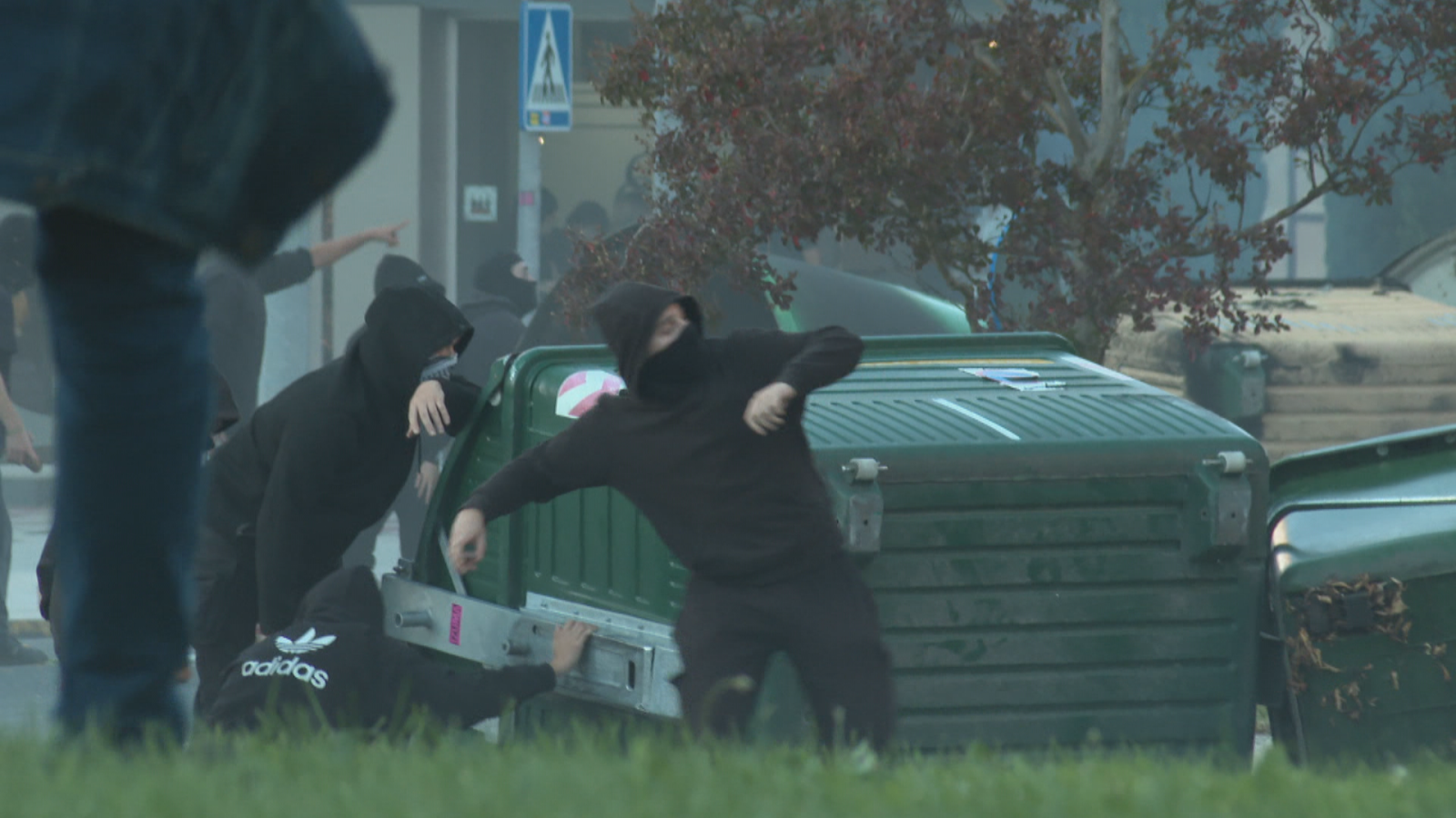 Varios manifestantes con el rostro cubierto y ropa oscura participan en disturbios urbanos junto a contenedores de basura que actúan como barricada.