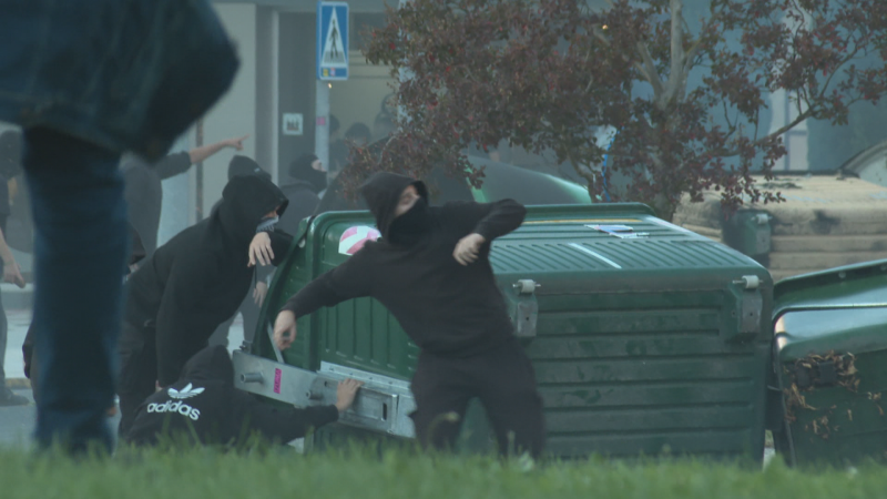 Varios manifestantes con el rostro cubierto y ropa oscura participan en disturbios urbanos junto a contenedores de basura que actúan como barricada.