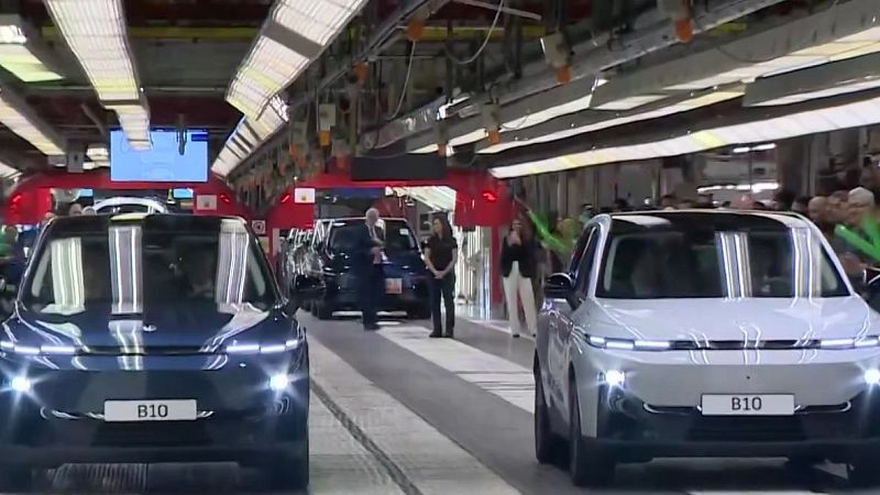 Interior de una fábrica de automóviles con dos SUV nuevos, uno azul y otro claro, y varias personas observando en la línea de ensamblaje.