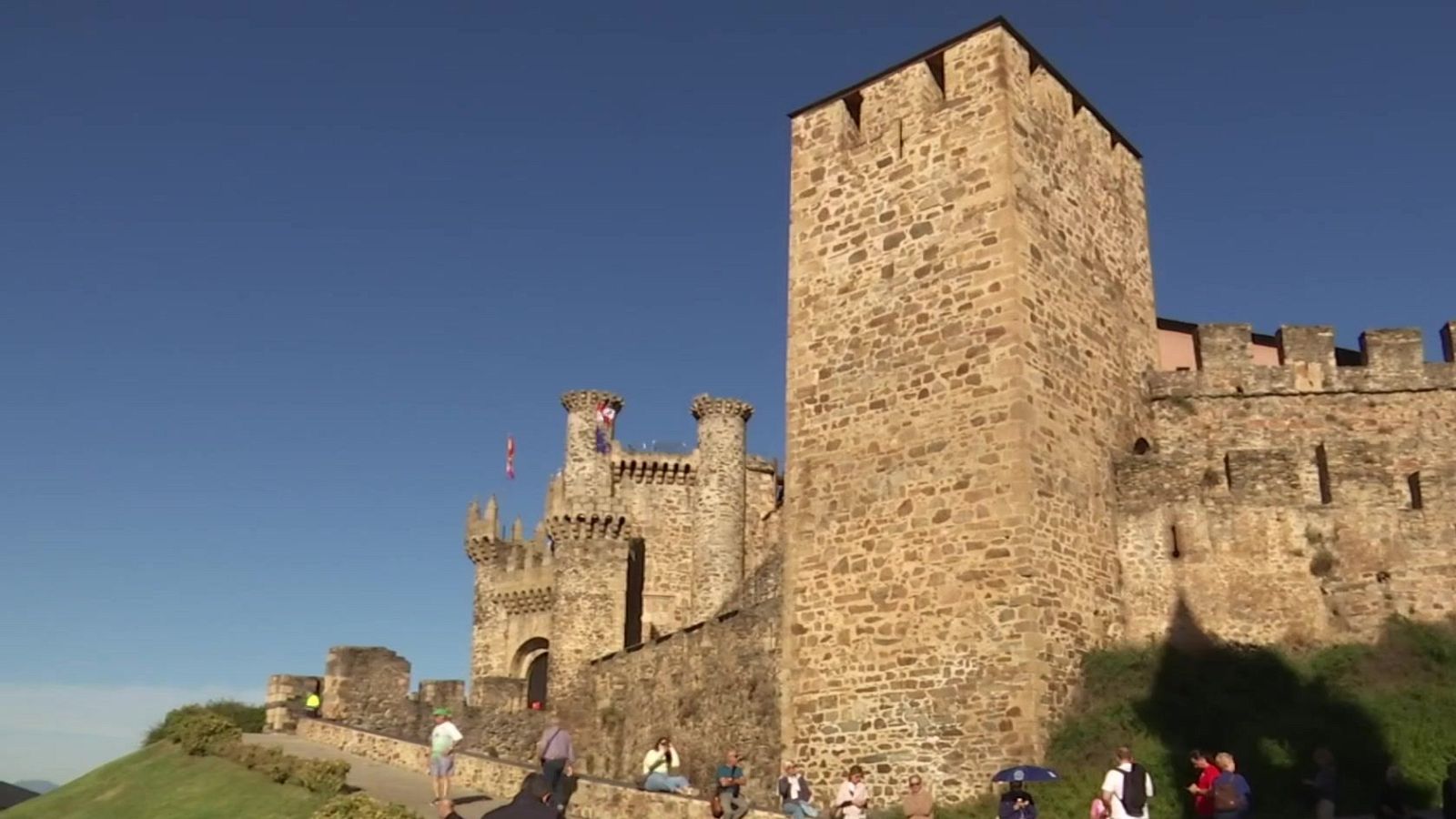 Vista exterior del Castillo de los Templarios en Ponferrada con su imponente torre, murallas almenadas y visitantes en el camino de acceso bajo un cielo azul.
