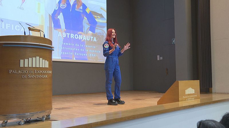 La astronauta de la ESA Sara García Alonso, con mono azul, habla en un auditorio ante una pantalla con su foto y la palabra ASTRONAUTA.