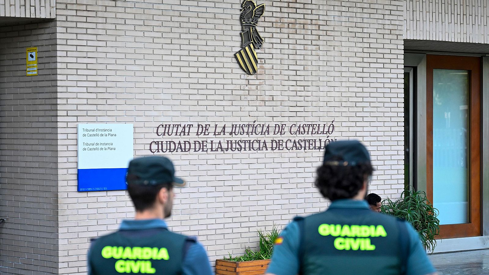 Dos agentes de la Guardia Civil de espaldas frente a la fachada y la puerta de acceso acristalada de la Ciudad de la Justicia.