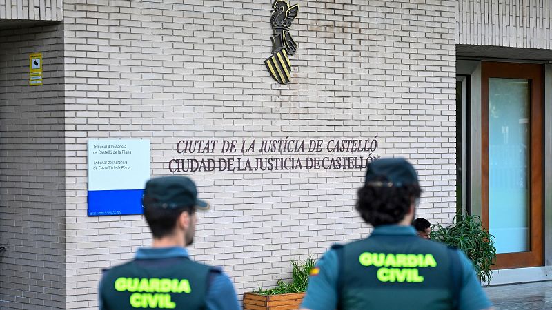 Dos agentes de la Guardia Civil de espaldas frente a la fachada y la puerta de acceso acristalada de la Ciudad de la Justicia.