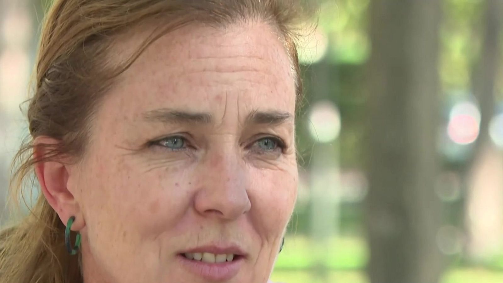 Primer plano de una mujer de mediana edad con pecas y cabello recogido, con expresión de estar hablando en un entorno exterior con vegetación desenfocada.