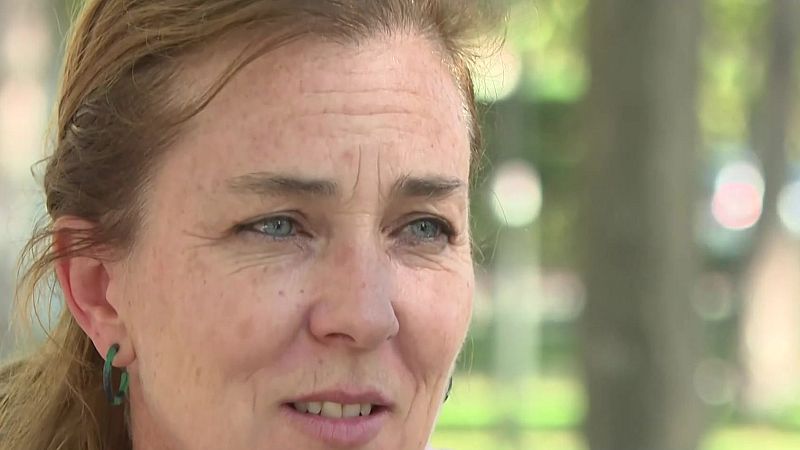 Primer plano de una mujer de mediana edad con pecas y cabello recogido, con expresión de estar hablando en un entorno exterior con vegetación desenfocada.