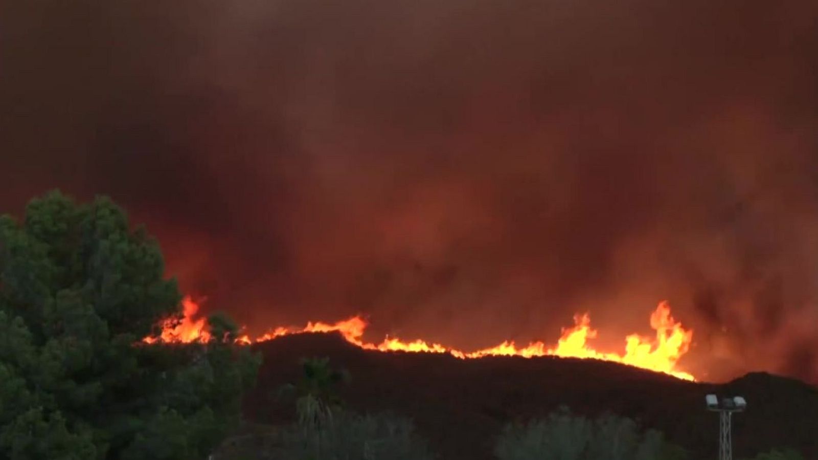 Incendio forestal nocturno en la cresta de una montaña con densas nubes de humo rojizo y una línea de fuego continuo devorando la vegetación.