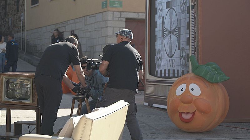 Plaza empedrada de un pueblo con el equipo técnico rodando junto a una gran figura de Naranjito y un televisor antiguo.
