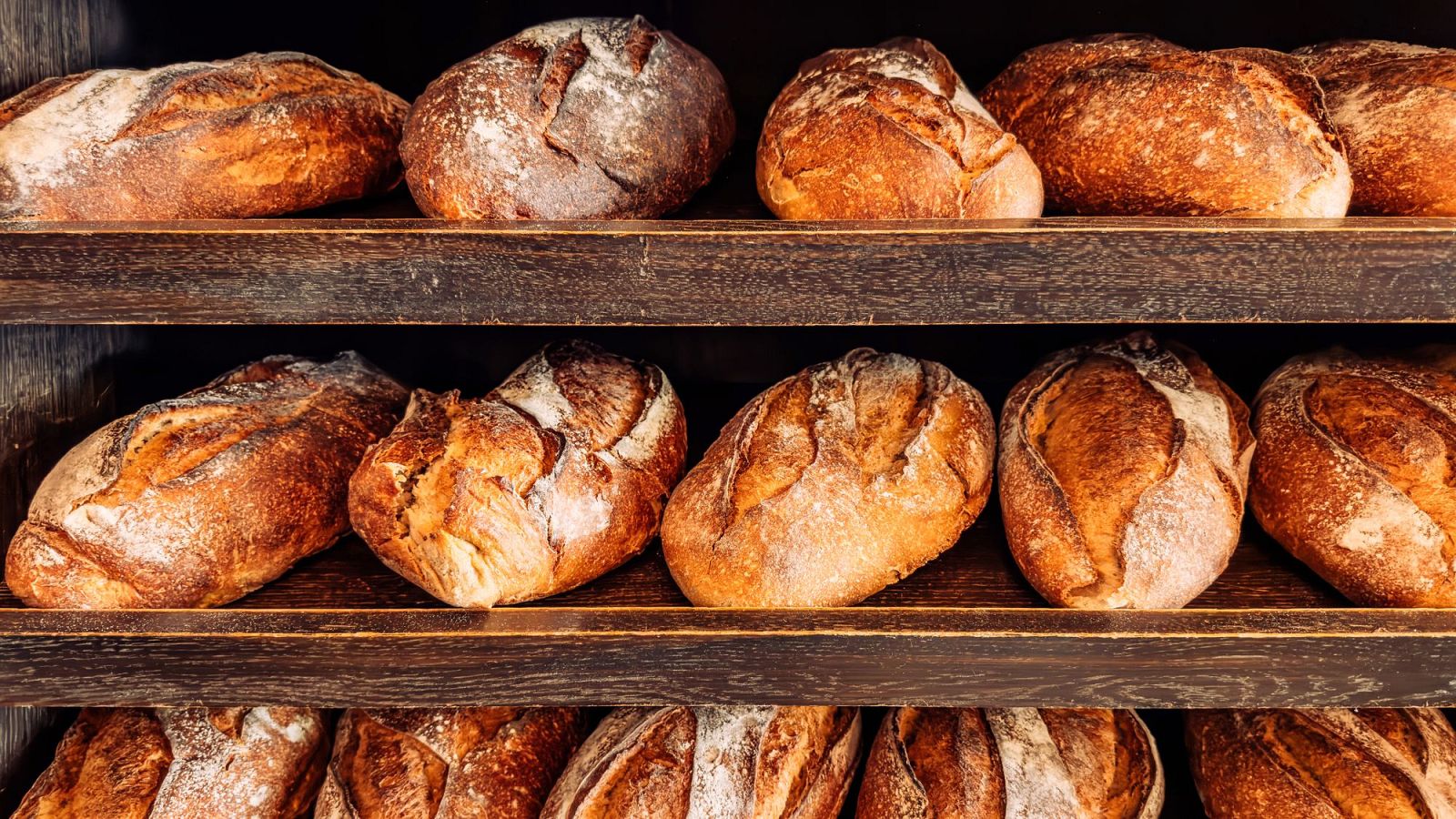 Expositor de panadería con tres estantes de madera oscura repletos de hogazas de pan rústico con cortezas tostadas.