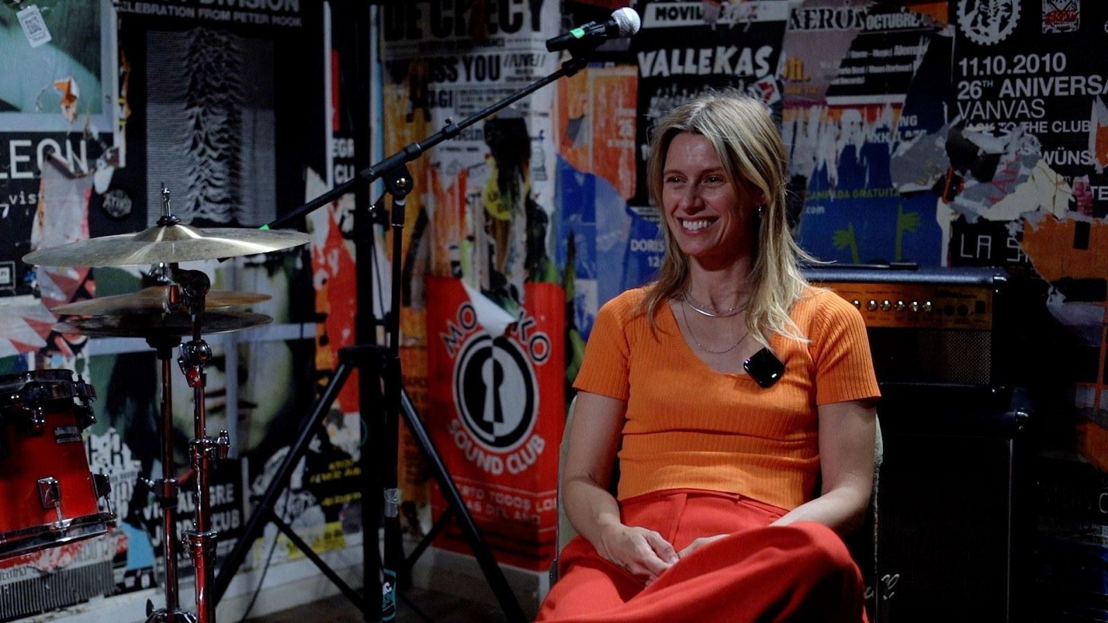 Mujer rubia sonriente con camiseta naranja y pantalón rojo sentada en un estudio musical decorado con carteles de conciertos y una batería.