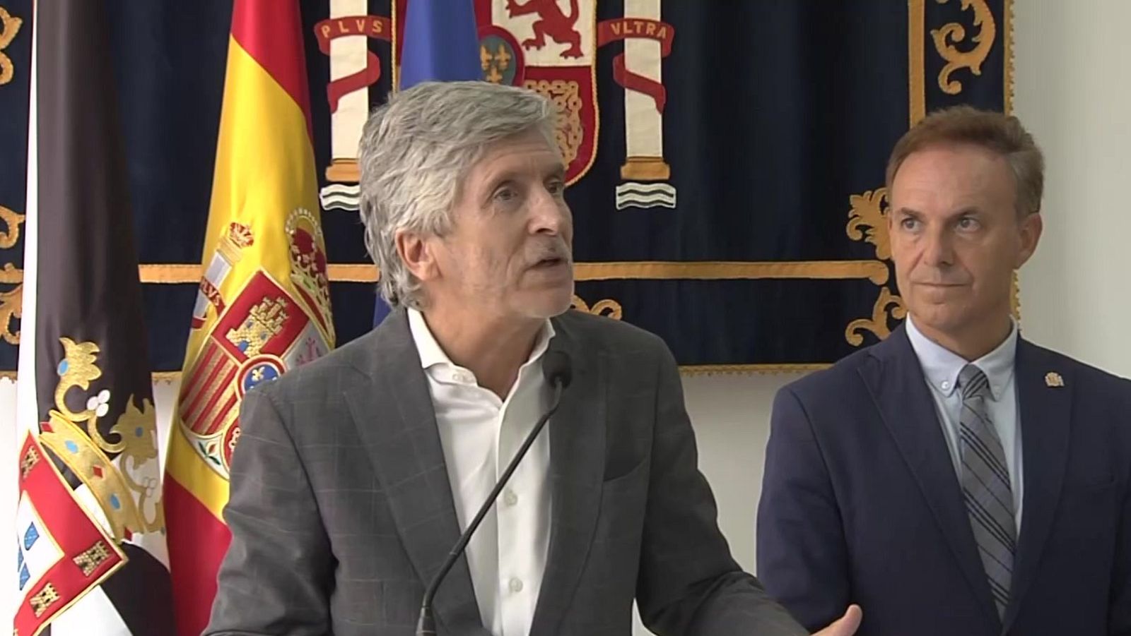 El ministro del Interior y el delegado del Gobierno en Ceuta comparecen juntos ante los medios en una rueda de prensa institucional con banderas de fondo.