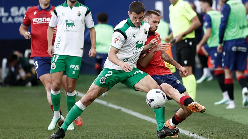 Disputa por el balón entre un futbolista con camiseta blanca y pantalón verde y un rival con camiseta roja.