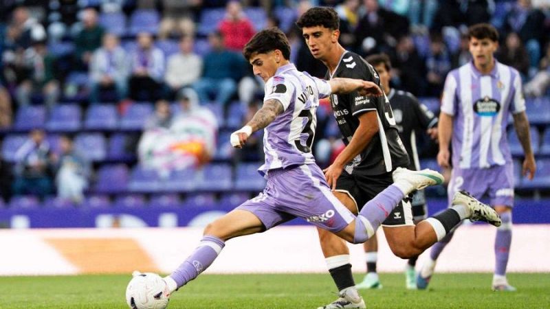 Un futbolista de un equipo blanquivioleta golpea el bal�n en el c�sped mientras un rival de negro presiona la jugada.