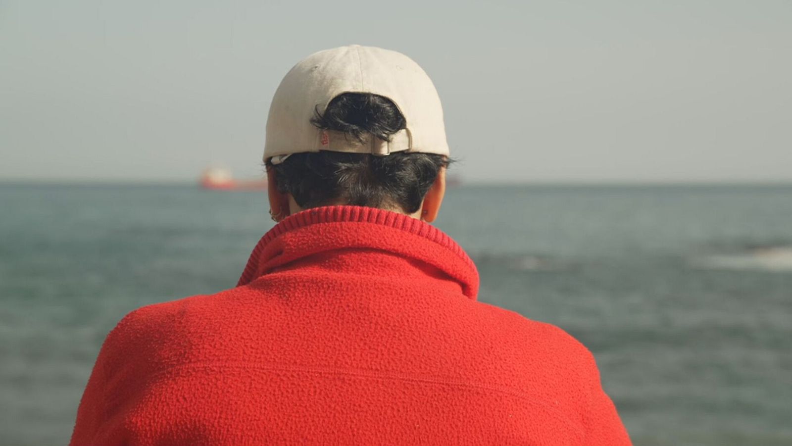 Persona de espaldas con forro polar rojo y gorra beige contempla el mar con una embarcación a lo lejos.