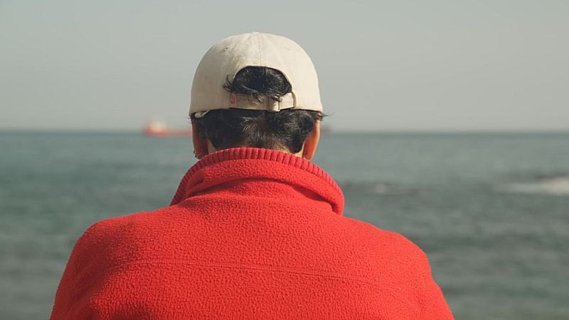 Persona de espaldas con forro polar rojo y gorra beige contempla el mar con una embarcación a lo lejos.