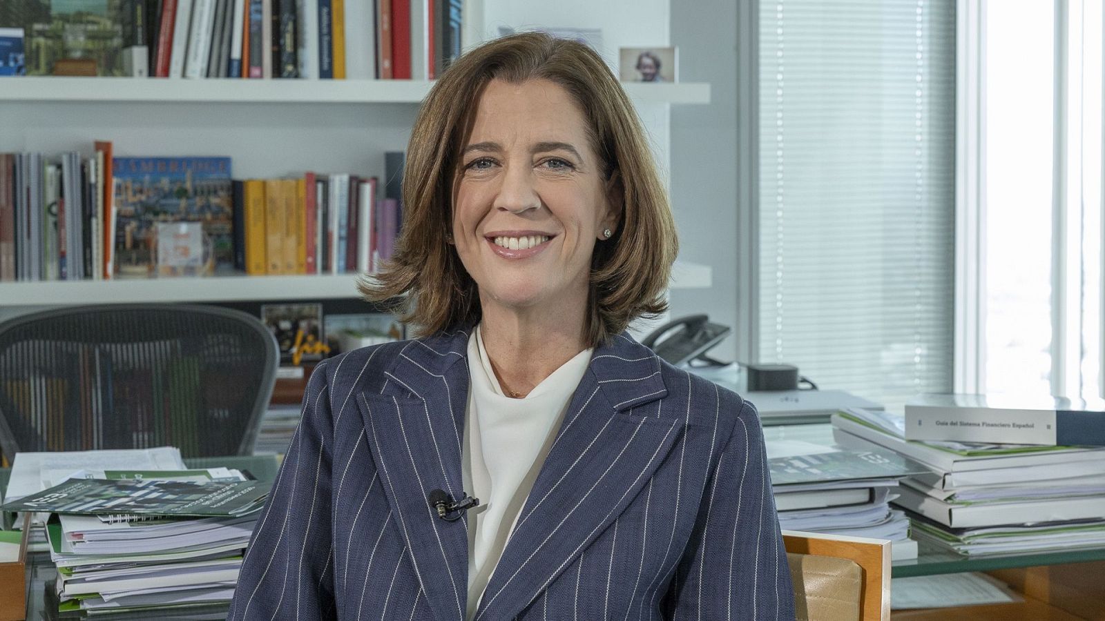 Alejandra Kindelán sonríe a cámara en su despacho profesional, vestida con chaqueta formal y rodeada de libros e informes.