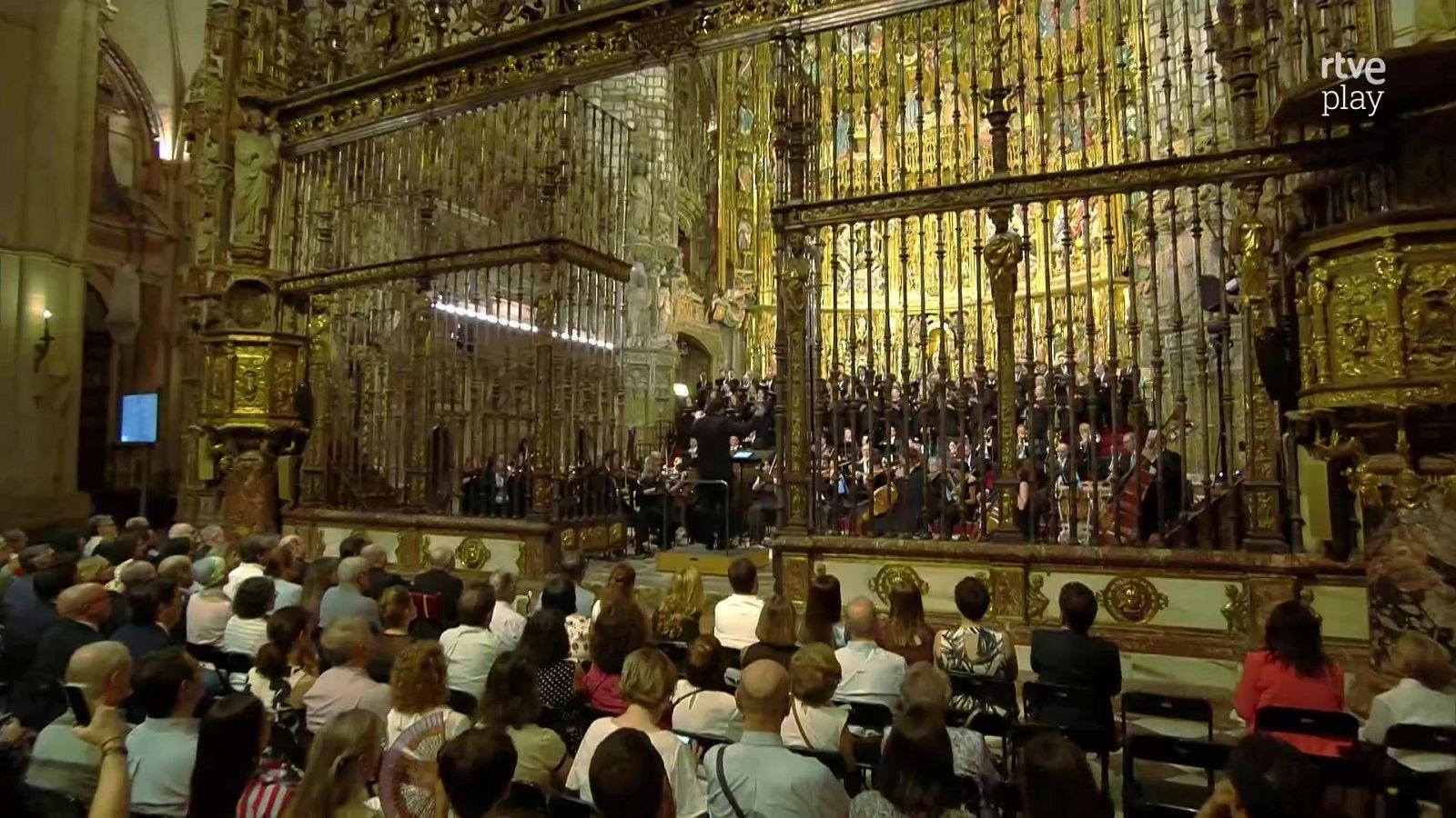 Catedral histórica durante un concierto de música clásica con orquesta, coro y público asistente de espaldas.