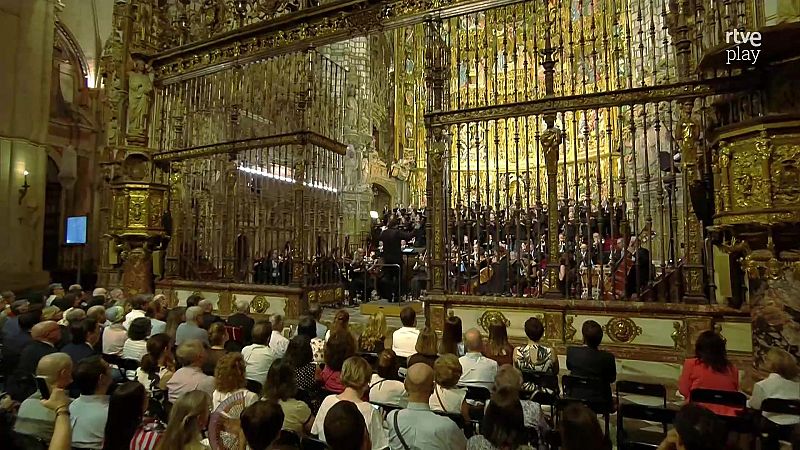 Catedral histórica durante un concierto de música clásica con orquesta, coro y público asistente de espaldas.